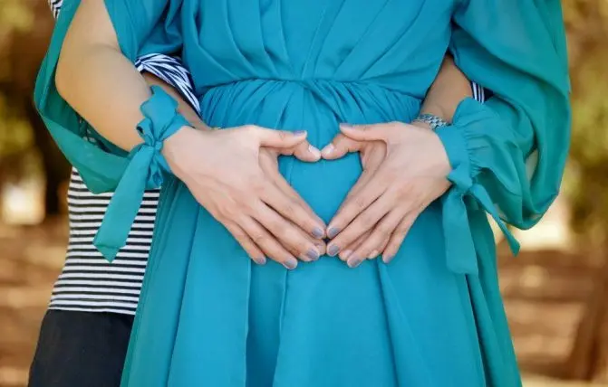 coppia forma con le mani un cuore sulla pancia della donna. La donna indossa un vestito color verde acqua